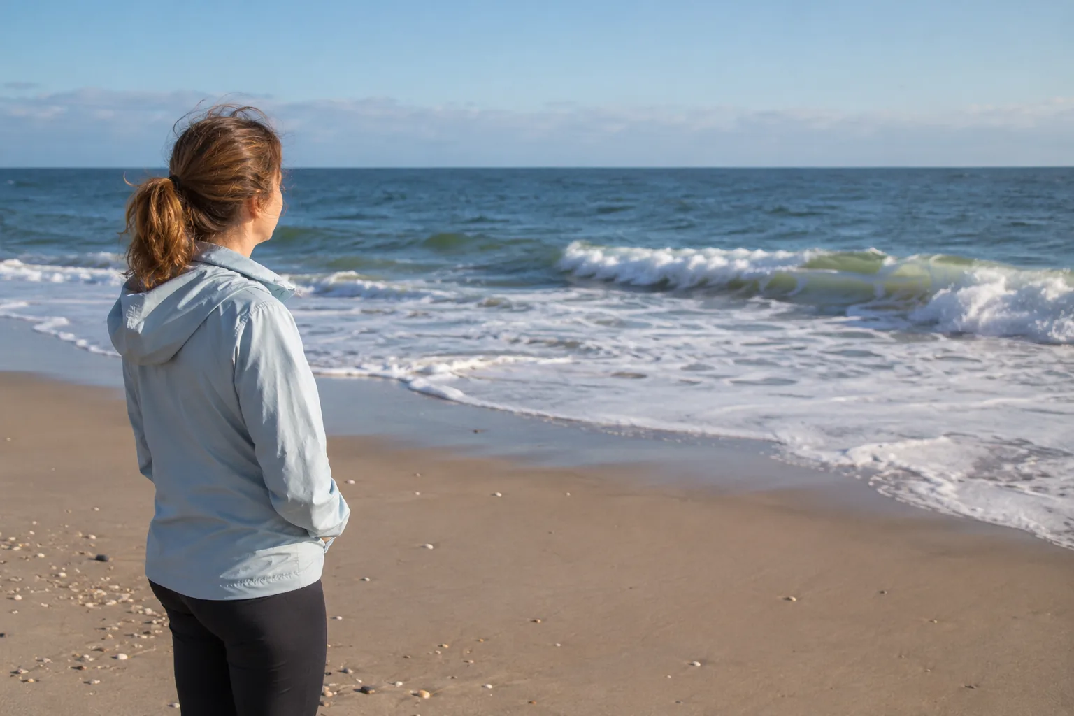 Visitor in a light jacket looking toward gentle Atlantic surf.